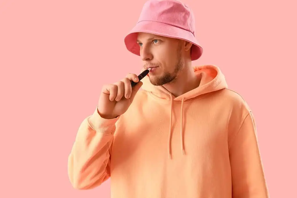 A man wearing a pink bucket hat smoking a cigarette on a pink background.
