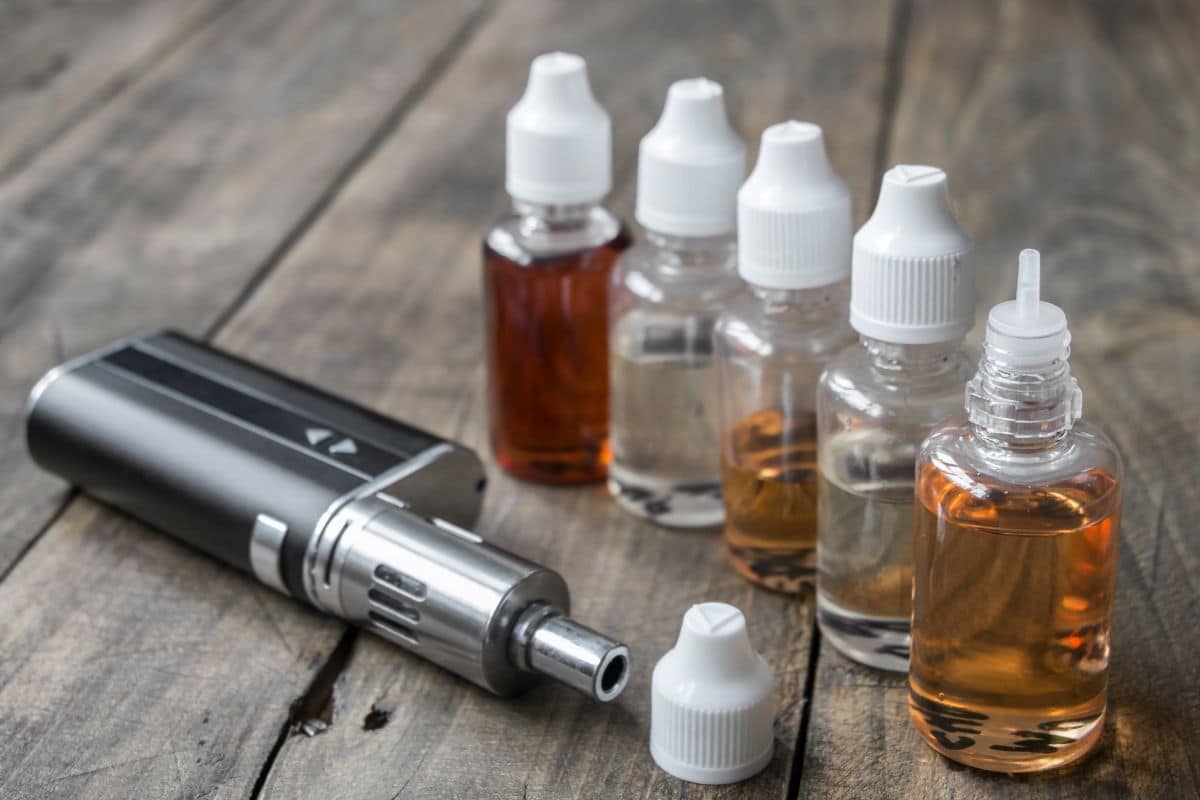 Top 8 Best Vapes Made in the USA: Quality and Innovation 4 A vape device lies on a wooden surface next to five plastic bottles of e-liquid in various shades of amber, each with a white cap. One bottle is uncapped, and the bottle caps are nearby.