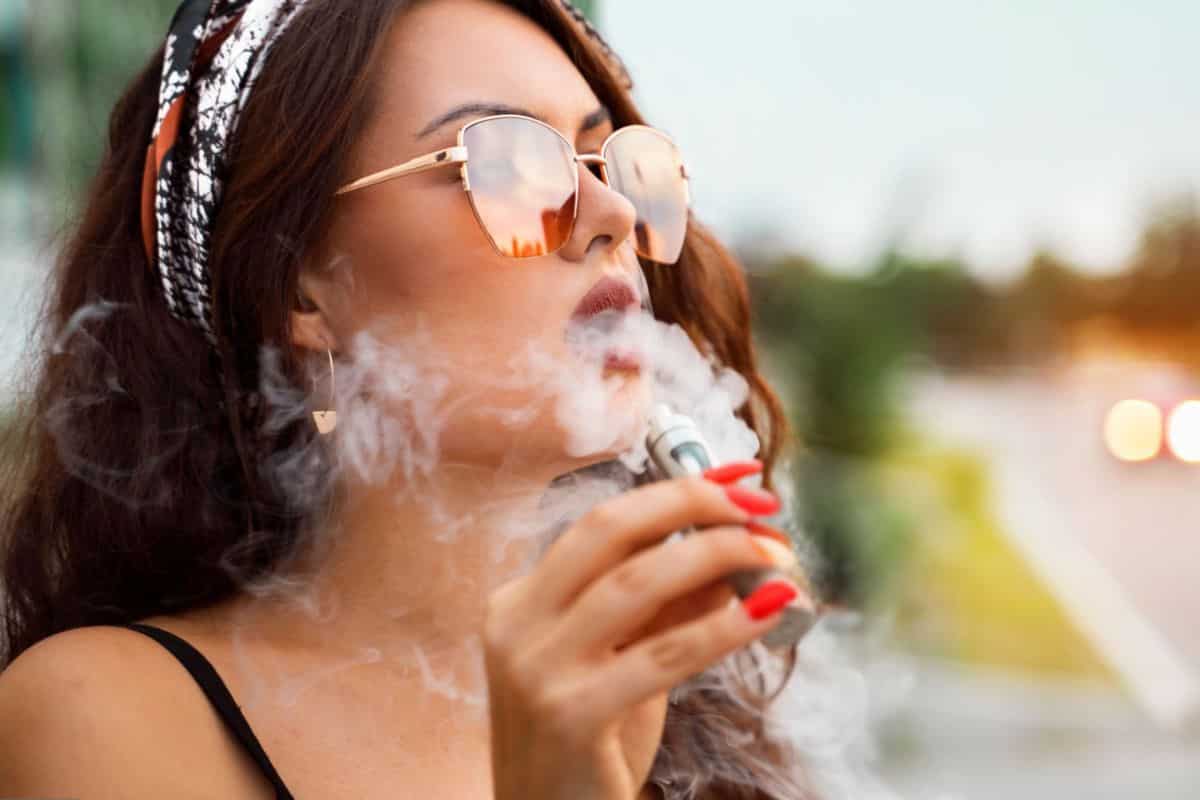 Top 8 Best Vapes Made in the USA: Quality and Innovation 3 A woman with long hair, wearing sunglasses and a patterned headband, exhales vapor while holding a vape device. She has red nail polish and is outdoors, surrounded by blurred greenery and lights.